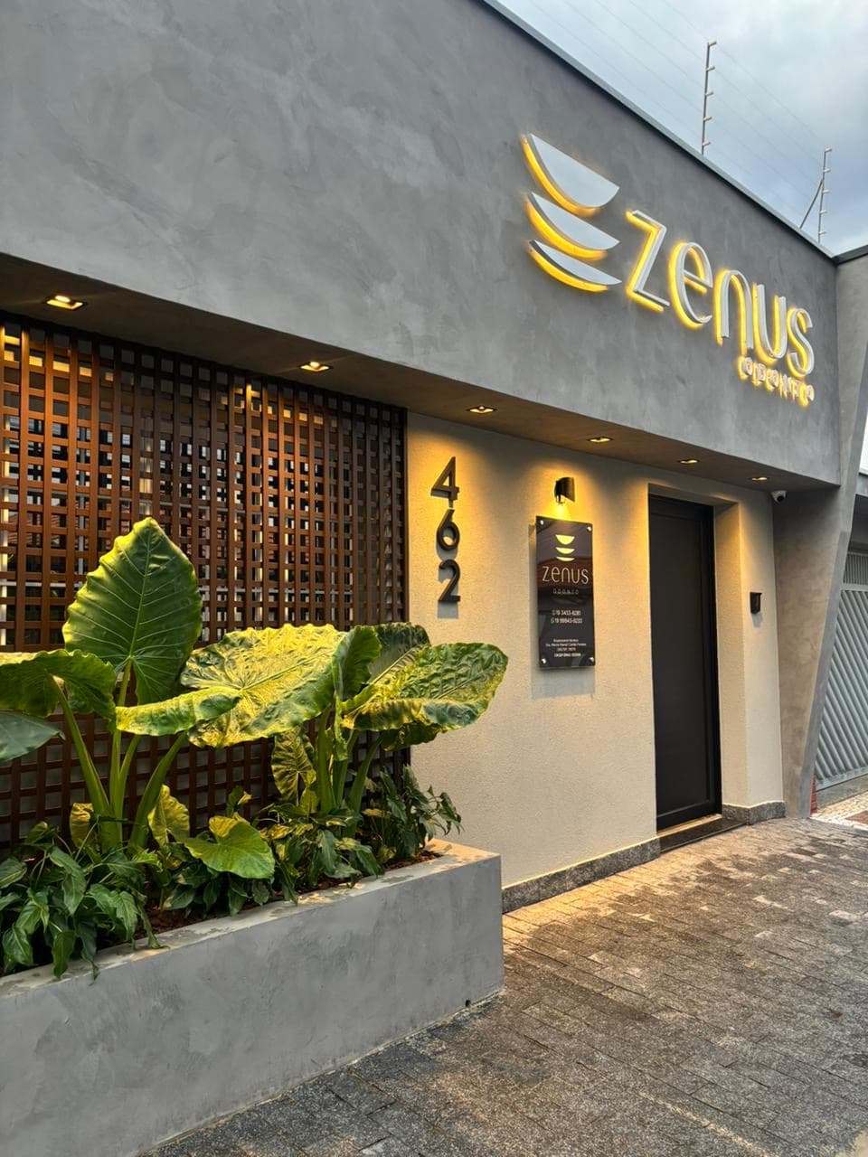 Fachada da Zenus Odonto ao anoitecer: o letreiro aceso, o número 462 e o muxarabi de madeira atrás das folhagens
