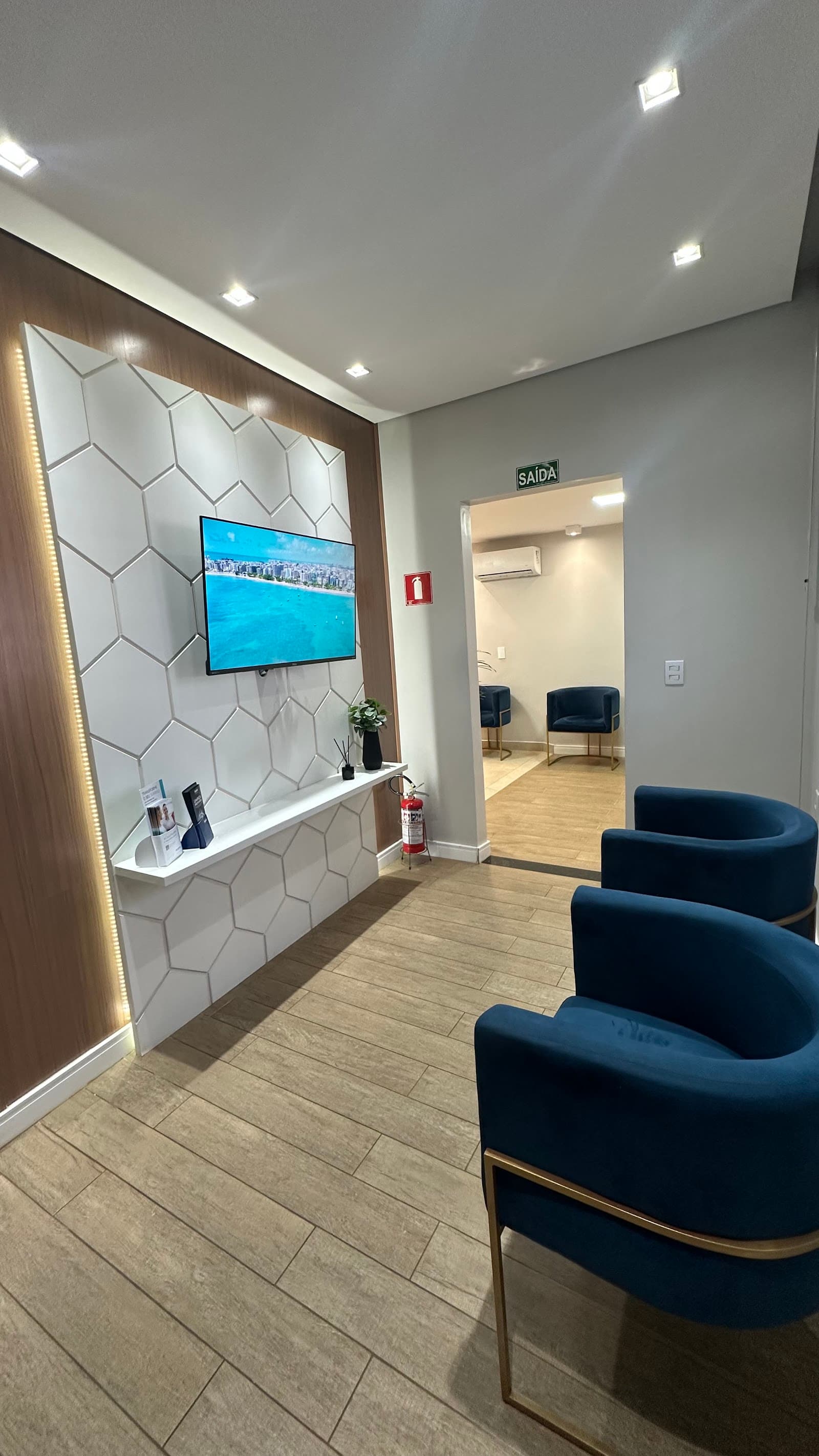 Sala de espera com poltronas de veludo azul, piso de madeira e painel branco de hexágonos com televisão